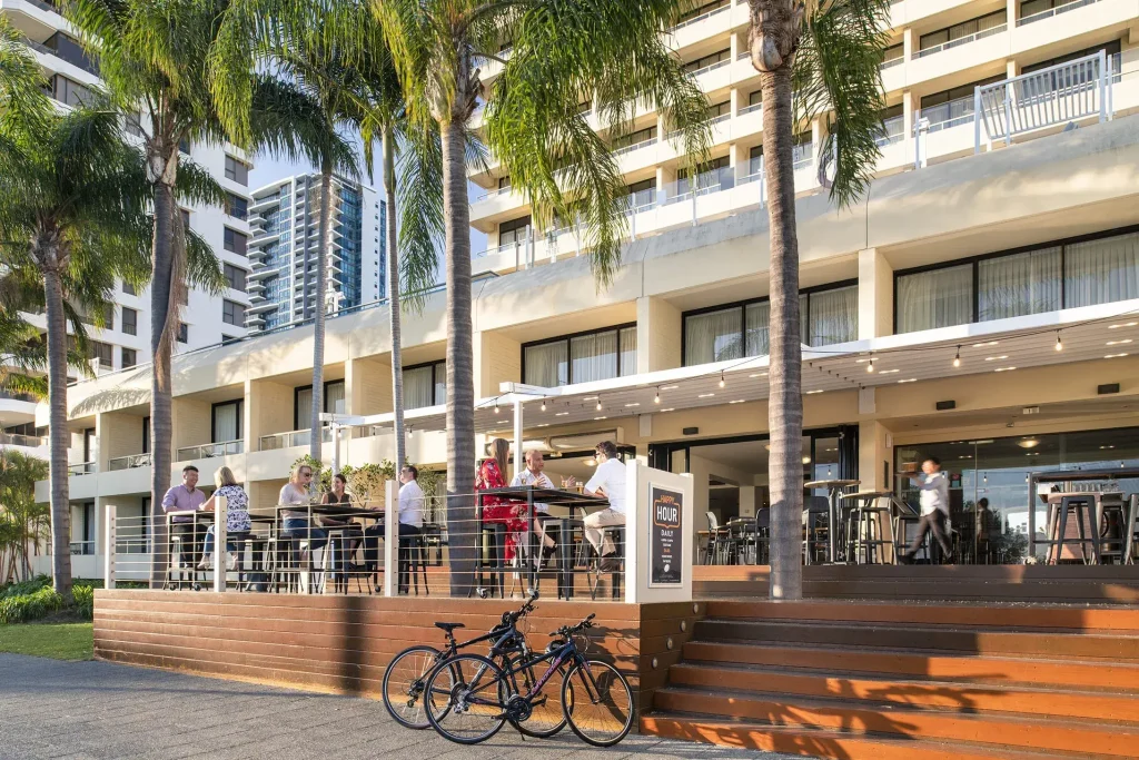 Outdoor hotel restaurant terrace with people dining at high tables under palm trees, set against a modern multi-story building, with bicycles parked in front.